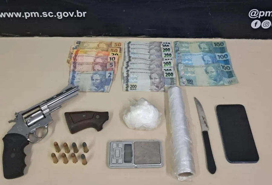 Itens apreendidos pela Polícia Militar em Criciúma dispostos sobre uma mesa: um revólver prata, oito munições, uma porção de cocaína, balança de precisão, rolo de plástico filme, faca, celular e diversas notas de Real (notas de 200, 100 e valores menores).
