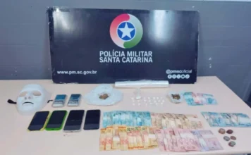 Orleans: Polícia fecha ponto de tráfico no São Jerônimo A imagem apresenta uma mesa branca com diversos materiais apreendidos pela Polícia Militar sobre ela. À esquerda, destaca-se uma máscara branca de rosto inteiro. Ao lado, quatro smartphones e duas balanças de precisão digitais. No centro, há porções de maconha e buchas de cocaína em plásticos, além de um rolo de plástico filme. À direita e na parte inferior, diversas notas de real (valores de 2 a 100) e moedas estão organizadas. Ao fundo, um painel preto exibe o brasão da Polícia Militar de Santa Catarina (PMSC), o site oficial e o perfil das redes sociais da instituição.