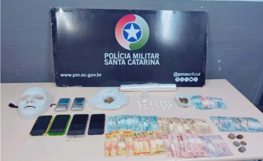 A imagem apresenta uma mesa branca com diversos materiais apreendidos pela Polícia Militar sobre ela. À esquerda, destaca-se uma máscara branca de rosto inteiro. Ao lado, quatro smartphones e duas balanças de precisão digitais. No centro, há porções de maconha e buchas de cocaína em plásticos, além de um rolo de plástico filme. À direita e na parte inferior, diversas notas de real (valores de 2 a 100) e moedas estão organizadas. Ao fundo, um painel preto exibe o brasão da Polícia Militar de Santa Catarina (PMSC), o site oficial e o perfil das redes sociais da instituição.