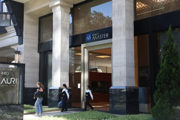 PF identifica R$ 2,2 bi ocultos por Vorcaro na Reag Fotografia da fachada do Banco Master, localizado na Avenida Faria Lima, em São Paulo. A imagem mostra a entrada principal do edifício, caracterizada por grandes colunas de pedra em estilo clássico e portas de vidro. No topo da entrada, vê-se o logotipo azul e a inscrição "Banco Master". No lado esquerdo, um totem indica o número 440 da via. Três mulheres caminham pela calçada em frente ao prédio, vestindo uniformes escuros e camisas brancas, sob a luz do dia.