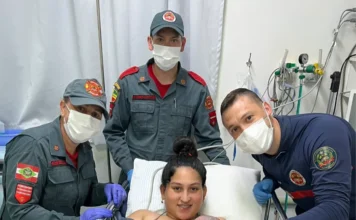 Partos assistidos pelos bombeiros em SC chegam a 111 em 2026 Uma jovem mãe sorridente, com aparelho e cabelo em um coque, está deitada em uma maca de hospital e segura seu bebê recém-nascido envolto em uma manta verde clara. Ela está coberta com um cobertor xadrez. Três socorristas do Corpo de Bombeiros Militar de Santa Catarina (CBMSC) estão ao redor dela, todos usando máscaras faciais brancas e luvas (roxas e azuis). À esquerda, uma bombeira de uniforme cinza com boné e o patch do CBMSC. Atrás da mãe, outro bombeiro com boné e uniforme cinza. À direita, um socorrista de uniforme azul escuro com detalhes vermelhos. Todos os socorristas estão sorrindo (visivelmente pelos olhos). O fundo mostra uma cortina de hospital branca e equipamentos médicos e tubos na parede. A foto transmite alegria e alívio após o parto.