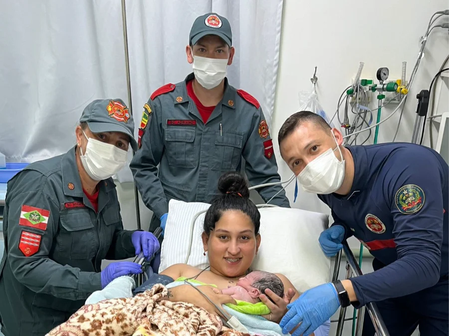 Uma jovem mãe sorridente, com aparelho e cabelo em um coque, está deitada em uma maca de hospital e segura seu bebê recém-nascido envolto em uma manta verde clara. Ela está coberta com um cobertor xadrez. Três socorristas do Corpo de Bombeiros Militar de Santa Catarina (CBMSC) estão ao redor dela, todos usando máscaras faciais brancas e luvas (roxas e azuis). À esquerda, uma bombeira de uniforme cinza com boné e o patch do CBMSC. Atrás da mãe, outro bombeiro com boné e uniforme cinza. À direita, um socorrista de uniforme azul escuro com detalhes vermelhos. Todos os socorristas estão sorrindo (visivelmente pelos olhos). O fundo mostra uma cortina de hospital branca e equipamentos médicos e tubos na parede. A foto transmite alegria e alívio após o parto.