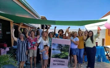 Passo de Torres promove evento para mulheres em Tapera Grupo de mulheres da comunidade de Tapera, em Passo de Torres, reunidas durante o evento de empoderamento e autoestima "O poder de ser você". As participantes aparecem sorridentes, algumas com os braços erguidos em sinal de força e união, posicionadas ao lado de um banner com a inscrição "Mulheres da Tapera" em um ambiente externo.