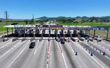 Pedágio na BR-101 Sul em SC sobe para R$ 3 nesta sexta Vista aérea detalhada da praça de pedágio da CCR ViaCosteira em Santa Catarina, mostrando cabines de cobrança manual e faixas de cobrança automática em uma rodovia movimentada, com cones e paisagem rural ao fundo sob céu azul.