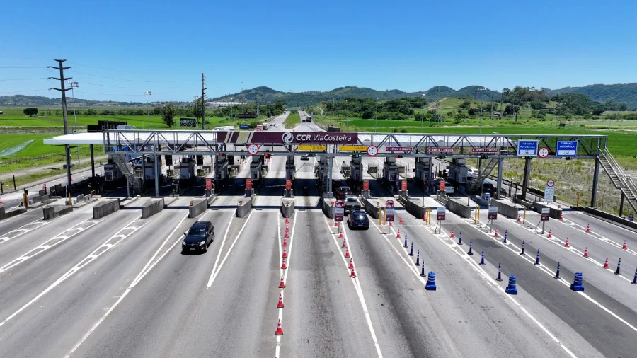 Vista aérea detalhada da praça de pedágio da CCR ViaCosteira em Santa Catarina, mostrando cabines de cobrança manual e faixas de cobrança automática em uma rodovia movimentada, com cones e paisagem rural ao fundo sob céu azul.