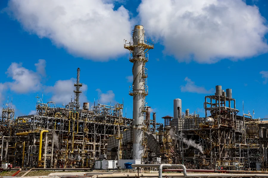 Vista detalhada de uma refinaria de petróleo com grandes torres de destilação, chaminés e uma complexa rede de tubulações sob um céu azul com nuvens brancas. A imagem ilustra a infraestrutura de refino que está no centro das atenções globais devido à volatilidade dos preços da commodity.