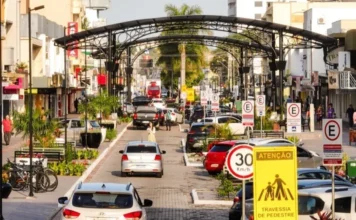 Pintura viária altera trânsito no calçadão de Araranguá Imagem da Avenida Getúlio Vargas em Araranguá, SC, mostrando a área do calçadão onde o trânsito será interrompido temporariamente para serviços de pintura viária. A cena inclui carros, o característico arco de metal preto sobre a via, bicicletas estacionadas, e sinalização de trânsito visível, como placas de 30 km/h e de atenção para pedestres.