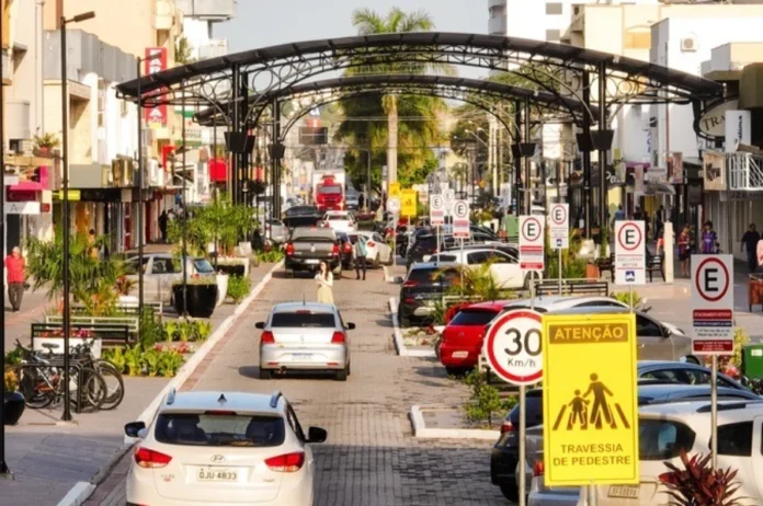 Pintura viária altera trânsito no calçadão de Araranguá Imagem da Avenida Getúlio Vargas em Araranguá, SC, mostrando a área do calçadão onde o trânsito será interrompido temporariamente para serviços de pintura viária. A cena inclui carros, o característico arco de metal preto sobre a via, bicicletas estacionadas, e sinalização de trânsito visível, como placas de 30 km/h e de atenção para pedestres.