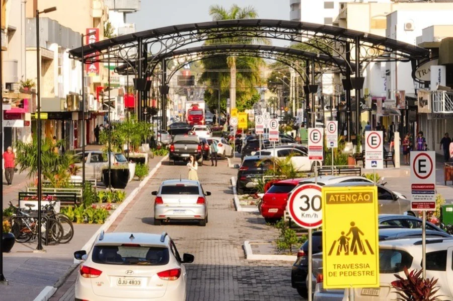 Imagem da Avenida Getúlio Vargas em Araranguá, SC, mostrando a área do calçadão onde o trânsito será interrompido temporariamente para serviços de pintura viária. A cena inclui carros, o característico arco de metal preto sobre a via, bicicletas estacionadas, e sinalização de trânsito visível, como placas de 30 km/h e de atenção para pedestres.
