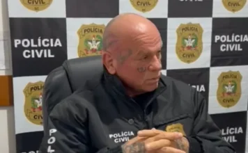 Polícia indicia trio por crime brutal dentro de presídio Um delegado homem calvo, com tatuagens no rosto (embaixo do olho) e nas mãos, sentado, com as mãos entrelaçadas. Ele veste uma jaqueta preta da Polícia Civil de Santa Catarina e está olhando para a direita. O fundo é composto por um painel de logotipos repetidos da Polícia Civil SC.