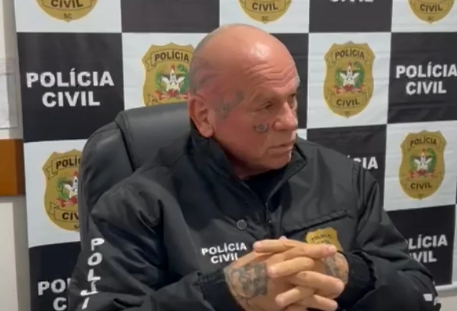 Um delegado homem calvo, com tatuagens no rosto (embaixo do olho) e nas mãos, sentado, com as mãos entrelaçadas. Ele veste uma jaqueta preta da Polícia Civil de Santa Catarina e está olhando para a direita. O fundo é composto por um painel de logotipos repetidos da Polícia Civil SC.