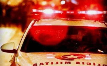 Polícia recupera veículo e persegue suspeito em área de mata Fotografia em plano médio da parte superior de uma viatura branca da Polícia Militar, com destaque para o giroflex aceso emitindo luzes vermelhas intensas. O capô do veículo exibe o brasão da corporação e a inscrição "POLÍCIA MILITAR" de forma espelhada. O fundo está desfocado, com luzes noturnas que criam um efeito de profundidade, reforçando a atmosfera de uma ocorrência policial em andamento.