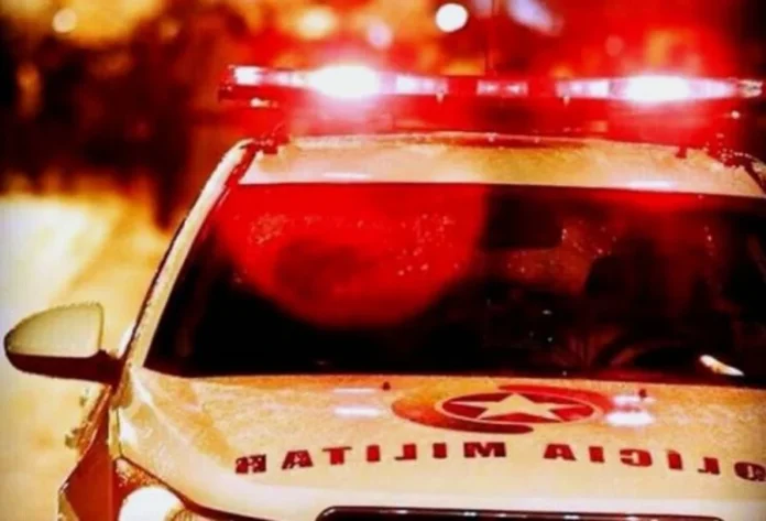 Polícia recupera veículo e persegue suspeito em área de mata Fotografia em plano médio da parte superior de uma viatura branca da Polícia Militar, com destaque para o giroflex aceso emitindo luzes vermelhas intensas. O capô do veículo exibe o brasão da corporação e a inscrição "POLÍCIA MILITAR" de forma espelhada. O fundo está desfocado, com luzes noturnas que criam um efeito de profundidade, reforçando a atmosfera de uma ocorrência policial em andamento.