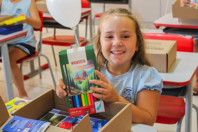 Praia Grande entrega kits de material escolar para 850 alunos da rede municipal pelo quinto ano consecutivo. Uma estudante da rede municipal de Praia Grande (SC), vestindo uniforme azul claro, sorri sentada em sua carteira escolar enquanto segura uma caixa de lápis de cor. À sua frente, há uma caixa de papelão aberta contendo outros itens do kit escolar, como apontador e cadernos. O ambiente ao fundo mostra uma sala de aula com cadeiras vermelhas e outros alunos.