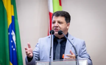 Praia Grande lança Expocanyon 2026 na Assembleia O prefeito de Praia Grande, Elisandro Fanica, discursa na tribuna da Assembleia Legislativa de Santa Catarina (Alesc) para convidar parlamentares e a sociedade para a Expocanyon 2026. Ele veste um paletó cinza claro, camisa escura e gravata, gesticulando enquanto fala aos microfones. Ao fundo, as bandeiras do Brasil e de Santa Catarina compõem o cenário da sessão.