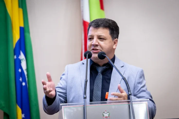 Praia Grande lança Expocanyon 2026 na Assembleia O prefeito de Praia Grande, Elisandro Fanica, discursa na tribuna da Assembleia Legislativa de Santa Catarina (Alesc) para convidar parlamentares e a sociedade para a Expocanyon 2026. Ele veste um paletó cinza claro, camisa escura e gravata, gesticulando enquanto fala aos microfones. Ao fundo, as bandeiras do Brasil e de Santa Catarina compõem o cenário da sessão.