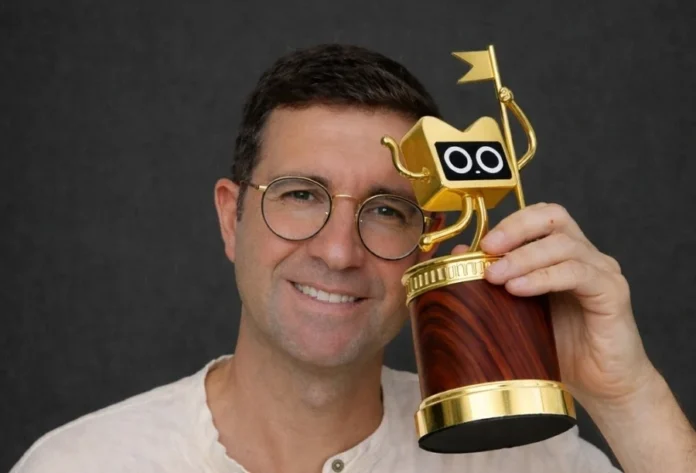 Professor da Unesc concorre a prêmio internacional de tecnologia Um plano médio de um homem de meia-idade, sorrindo e olhando para a câmera, usando óculos redondos de armação dourada e uma camisa de linho branco. Ele está segurando um troféu dourado exclusivo e detalhado na mão direita contra um fundo cinza escuro. O troféu tem uma base cilíndrica de madeira de grão escuro. No topo, há uma escultura de robô dourado estilizado com um corpo em forma de caixa, "olhos" de robô mecânico (círculos brancos e pretos) e um pequeno braço segurando uma haste com uma pequena bandeira. O homem tem cabelos castanhos curtos e uma expressão de alegria e realização.