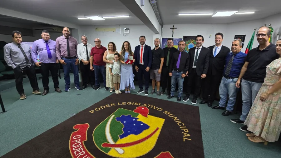 Professora Juliana Martins Vargas segura a Medalha Padre João Reitz ao lado de vereadores e familiares no plenário da Câmara Municipal de Sombrio durante sessão de homenagem por ato de bravura.