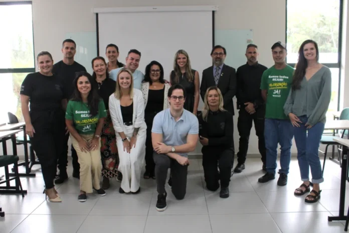 Projeto busca reduzir reincidência de violência doméstica Uma fotografia de grupo colorida, em plano médio, de 14 pessoas posando em uma sala de aula ou auditório na Unesc. Elas estão organizadas em duas filas: seis pessoas agachadas na frente e oito de pé atrás, todas voltadas para a câmera. No centro, agachado, um homem de óculos e camisa polo azul claro. À esquerda dele, uma mulher agachada veste uma camiseta verde com a inscrição "Somos todos SELURE. Valorização JÁ!". À direita dele, outra mulher agachada veste roupas pretas. As pessoas de pé atrás usam roupas variadas, incluindo ternos, uniformes pretos e roupas casuais. O fundo é uma sala limpa com grandes janelas que trazem luz natural, uma tela de projeção branca vazia e mesas de estudo dobradas à esquerda. O chão é de piso frio.
