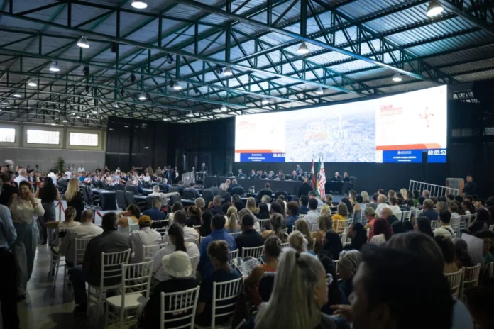 Próxima Alesc Itinerante será em Araranguá em maio Grande público assiste a sessão da Alesc Itinerante em Curitibanos, Santa Catarina, com deputado Julio Garcia discursando no palco. A foto mostra cadeiras brancas e um grande painel de LED ao fundo com a marca do programa.