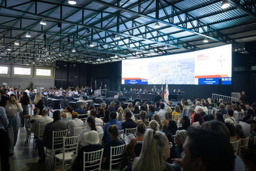 Grande público assiste a sessão da Alesc Itinerante em Curitibanos, Santa Catarina, com deputado Julio Garcia discursando no palco. A foto mostra cadeiras brancas e um grande painel de LED ao fundo com a marca do programa.