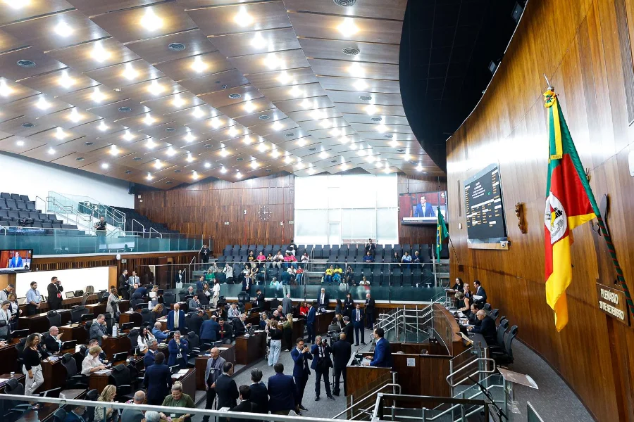 Foto ampla do interior do Plenário 20 de Setembro da Assembleia Legislativa do Rio Grande do Sul (ALRS), em Porto Alegre. O espaço é caracterizado por teto e paredes de madeira, e múltiplas luzes embutidas. O nível inferior está movimentado com dezenas de deputados estaduais e servidores sentados em mesas e em pé, durante uma sessão de votação. Ao fundo, uma galeria de público em anfiteatro com assentos vazios e algumas pessoas observando. À direita, destacam-se as bandeiras do Brasil e do Rio Grande do Sul, penduradas ao lado de um grande painel eletrônico de votação com o brasão estadual e uma tela de vídeo maior mostrando uma transmissão ao vivo. O ambiente é formal e movimentado.