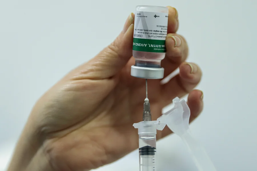 Mãos de profissional de saúde preparando dose de vacina inativada contra gripe Influenza A(H1N1) e A(H3N2) com seringa e frasco, com rótulo visível do SUS.