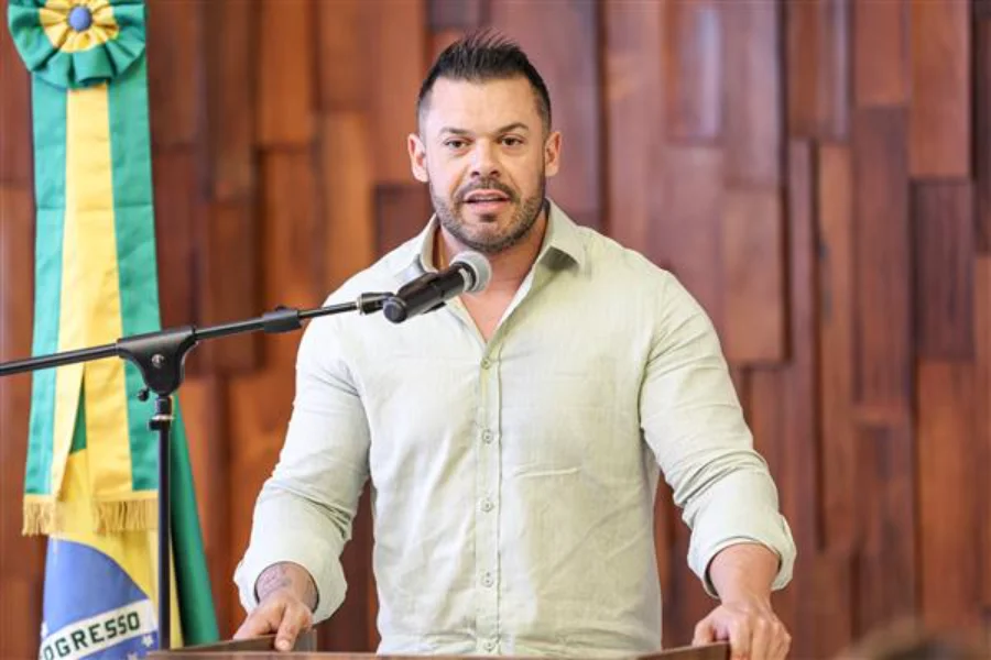 Fotografia de médio plano do deputado estadual Kaká D’Ávila (Podemos) durante pronunciamento na Assembleia Legislativa do Rio Grande do Sul. O parlamentar, que tem cabelo curto escuro e barba por fazer, veste uma camisa de linho verde-clara e fala ao microfone posicionado em um púlpito de madeira. Ele mantém as mãos apoiadas nas bordas do púlpito enquanto olha para a frente. À esquerda da imagem, vê-se parte da bandeira do Brasil. O fundo é composto por painéis de madeira verticais.
