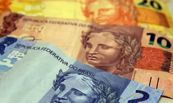 Receita Federal divulga regras do Imposto de Renda 2026 Um close-up de várias cédulas de dinheiro Real brasileiro sobrepostas. No primeiro plano, com foco nítido, a efígie da República em uma cédula azul (provavelmente de 100 reais) sobrepõe-se a uma nota laranja-amarronzada com o número '10' visível, referente a dez reais. O fundo é composto por outras notas desfocadas de cores variadas (laranja e amarelo), sugerindo mais denominações. A imagem destaca a textura do papel e os detalhes das gravuras, incluindo a efígie da República e a frase "REPÚBLICA FEDERATIVA DO BRASIL".