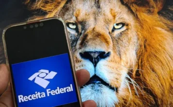 Receita Federal recebe 450 mil declarações no 1º dia Close de mão segurando smartphone com logotipo da Receita Federal em primeiro plano, tendo ao fundo a imagem de um leão em uma tela de computador, simbolizando o Imposto de Renda (IRPF).