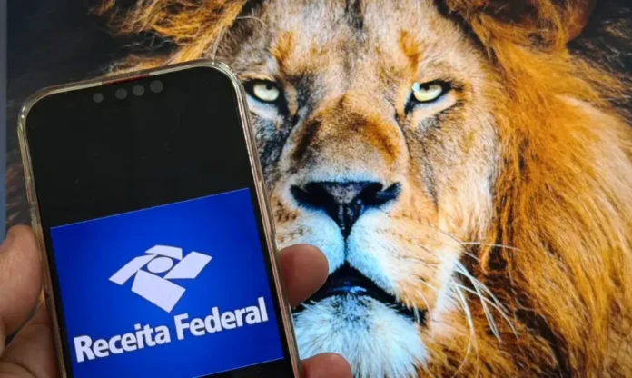 Receita Federal recebe 450 mil declarações no 1º dia (1) Close de mão segurando smartphone com logotipo da Receita Federal em primeiro plano, tendo ao fundo a imagem de um leão em uma tela de computador, simbolizando o Imposto de Renda (IRPF).