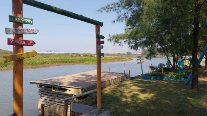 Reeducandos realizam manutenção em espaços de Araranguá Uma vista de Araranguá na comunidade de Ilhas com um deck de madeira em primeiro plano. Uma placa rústica de madeira com setas coloridas exibe frases inspiradoras como "Crie Ondas", "Coisas Boas", "Porque Elas" e "Amor". Ao fundo, em um gramado verde, reeducandos do Projeto Recomeçar realizam serviços de manutenção e limpeza próximos ao rio e árvores.