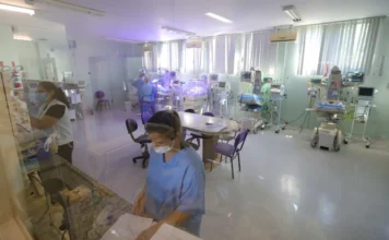 Residência em saúde em SC: inscrições encerram nesta quarta Profissionais de saúde vestindo uniformes azuis e máscaras protetoras trabalham em uma unidade hospitalar equipada com incubadoras neonatais e monitores de alta tecnologia. A imagem, capturada através de um vidro, mostra a rotina de assistência em uma ala de tratamento intensivo, representando o ambiente de atuação prática dos residentes na rede pública de Santa Catarina.