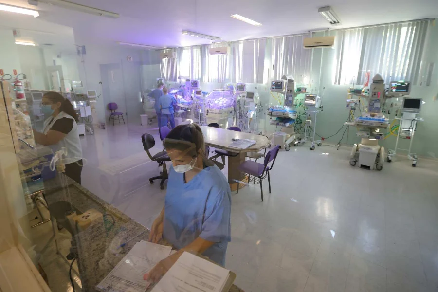 Residência em saúde em SC inscrições encerram nesta quarta Profissionais de saúde vestindo uniformes azuis e máscaras protetoras trabalham em uma unidade hospitalar equipada com incubadoras neonatais e monitores de alta tecnologia. A imagem, capturada através de um vidro, mostra a rotina de assistência em uma ala de tratamento intensivo, representando o ambiente de atuação prática dos residentes na rede pública de Santa Catarina.