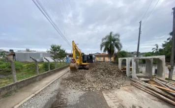 Rua Timbé do Sul é bloqueada para obras em Araranguá Foto de rua mostrando obras de infraestrutura em andamento sob um céu nublado. Uma escavadeira amarela está posicionada no centro de uma via inacabada, operando sobre um grande monte de pedras britadas e detritos de asfalto. À direita, grandes seções pré-moldadas de tubos de bueiro de concreto estão empilhadas, ladeadas por toras de madeira. O fundo exibe casas residenciais simples de um andar e uma palmeira proeminente, com postes de energia e fiação elétrica visíveis. Um trabalhador é visível na cabine da escavadeira.