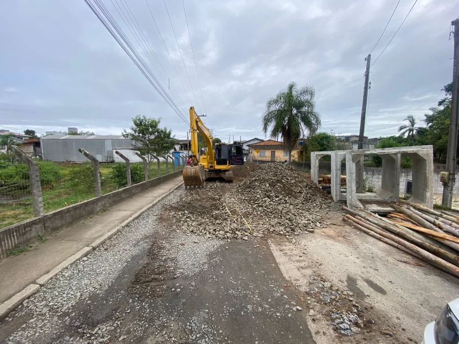 Foto de rua mostrando obras de infraestrutura em andamento sob um céu nublado. Uma escavadeira amarela está posicionada no centro de uma via inacabada, operando sobre um grande monte de pedras britadas e detritos de asfalto. À direita, grandes seções pré-moldadas de tubos de bueiro de concreto estão empilhadas, ladeadas por toras de madeira. O fundo exibe casas residenciais simples de um andar e uma palmeira proeminente, com postes de energia e fiação elétrica visíveis. Um trabalhador é visível na cabine da escavadeira.