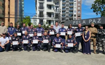 SAMU SC forma primeira turma de condutores de motolância Fotografia em plano aberto mostra um grupo de aproximadamente 25 pessoas posando para uma foto oficial em um pátio externo sob céu claro. A maioria dos presentes veste o uniforme azul-marinho do SAMU com detalhes em laranja e segura certificados brancos. Ao lado dos formandos, encontram-se policiais militares com fardas camufladas e cáqui, além de autoridades civis. Ao fundo, destaca-se uma estrutura de madeira com a inscrição parcial "PELA PAZ E PE" e edifícios residenciais.