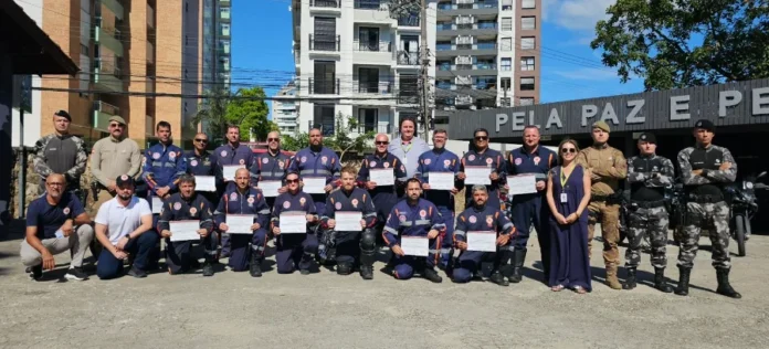 SAMU SC forma primeira turma de condutores de motolância Fotografia em plano aberto mostra um grupo de aproximadamente 25 pessoas posando para uma foto oficial em um pátio externo sob céu claro. A maioria dos presentes veste o uniforme azul-marinho do SAMU com detalhes em laranja e segura certificados brancos. Ao lado dos formandos, encontram-se policiais militares com fardas camufladas e cáqui, além de autoridades civis. Ao fundo, destaca-se uma estrutura de madeira com a inscrição parcial "PELA PAZ E PE" e edifícios residenciais.