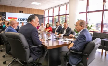 SC: Novo piso regional é entregue ao governador Governador Jorginho Mello e o presidente da FIESC, Gilberto Seleme, lideram reunião oficial no Centro Administrativo de Santa Catarina para a entrega da proposta de reajuste do piso salarial regional. Outros representantes de trabalhadores e empregadores estão sentados ao redor de uma grande mesa de conferência de madeira. O Governador está sentado na cabeceira oposta da mesa, conversando com Gilberto Seleme (visível em primeiro plano, de perfil à direita). O logotipo 'GOVERNO DE SANTA CATARINA' é visível em uma tela ao fundo, e grandes janelas mostram a vista de Florianópolis.