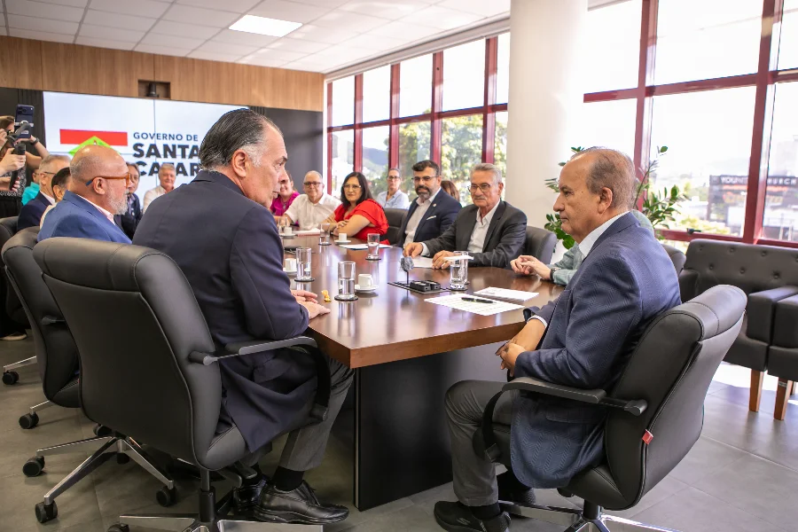 Governador Jorginho Mello e o presidente da FIESC, Gilberto Seleme, lideram reunião oficial no Centro Administrativo de Santa Catarina para a entrega da proposta de reajuste do piso salarial regional. Outros representantes de trabalhadores e empregadores estão sentados ao redor de uma grande mesa de conferência de madeira. O Governador está sentado na cabeceira oposta da mesa, conversando com Gilberto Seleme (visível em primeiro plano, de perfil à direita). O logotipo 'GOVERNO DE SANTA CATARINA' é visível em uma tela ao fundo, e grandes janelas mostram a vista de Florianópolis.