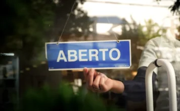 SC abre adesão ao Recupera Mais com descontos de até 95% Mão segura placa azul com a palavra "Aberto" escrita em branco em uma porta de vidro, simbolizando a retomada do comércio e o empreendedorismo em Santa Catarina.