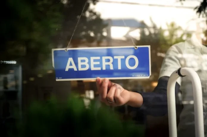 SC abre adesão ao Recupera Mais com descontos de até 95% Mão segura placa azul com a palavra "Aberto" escrita em branco em uma porta de vidro, simbolizando a retomada do comércio e o empreendedorismo em Santa Catarina.