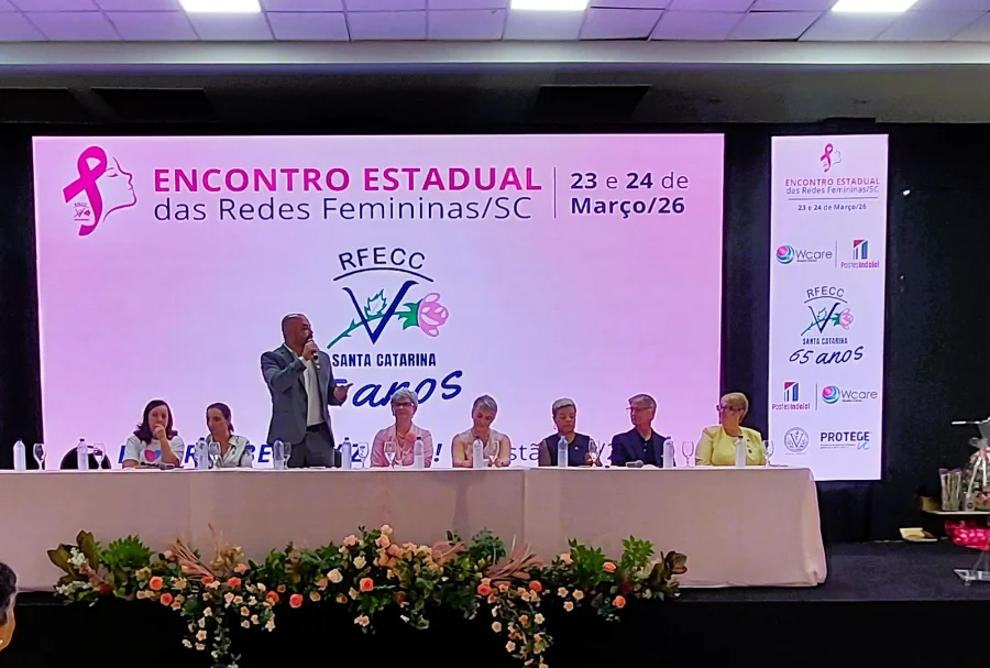 Foto do Secretário de Estado da Saúde de Santa Catarina, Diogo Demarchi, discursando no Encontro Estadual das Redes Femininas de Combate ao Câncer (RFECC SC). No painel de LED ao fundo, lê-se 'Encontro Estadual das Redes Femininas/SC' e 'RFECC Santa Catarina 65 Anos'. Pessoas estão sentadas na mesa principal.