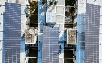 SC aprova energia solar para hospitais filantrópicos Vista aérea de drone de uma grande instalação de módulos de energia solar fotovoltaica em um telhado industrial de metal. A imagem mostra três seções principais de painéis solares azul-escuros cobrindo a estrutura do telhado, com equipamentos de ventilação e claraboias visíveis.