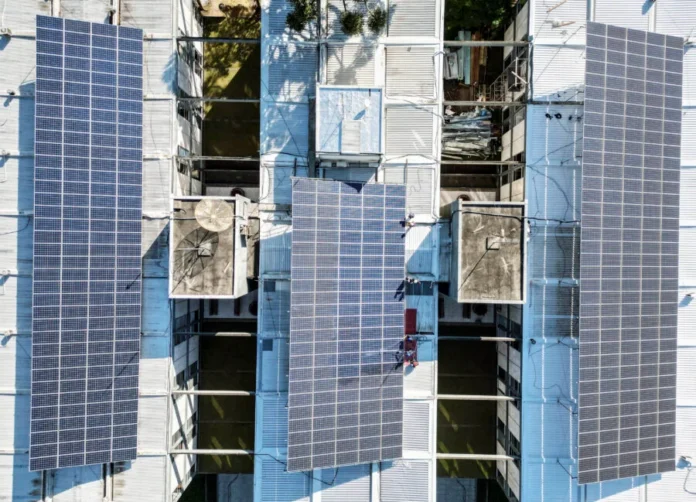 SC aprova energia solar para hospitais filantrópicos Vista aérea de drone de uma grande instalação de módulos de energia solar fotovoltaica em um telhado industrial de metal. A imagem mostra três seções principais de painéis solares azul-escuros cobrindo a estrutura do telhado, com equipamentos de ventilação e claraboias visíveis.