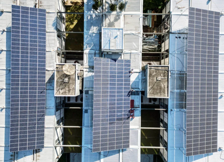 Vista aérea de drone de uma grande instalação de módulos de energia solar fotovoltaica em um telhado industrial de metal. A imagem mostra três seções principais de painéis solares azul-escuros cobrindo a estrutura do telhado, com equipamentos de ventilação e claraboias visíveis.