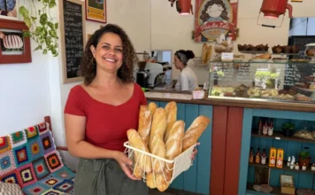 SC atinge 490 mil empresas lideradas por mulheres Uma foto de plano médio de Leslie Araújo, uma mulher com cabelos cacheados e vestindo uma blusa vermelha de decote ombro a ombro, sorrindo amplamente e segurando com as duas mãos uma cesta de arame branca cheia de várias baguetes douradas e artesanais de pão. Ela está em pé no interior de uma padaria aconchegante e rústica, com um balcão de serviço azul e madeira ao fundo. Atrás do balcão, outra mulher com um lenço na cabeça trabalha perto de uma vitrine de vidro cheia de doces e pães. Uma lousa de menu escrita à mão em português e uma placa redonda ilustrada que diz "Pão da Leli" são visíveis acima do balcão. À esquerda, prateleiras de madeira com fotos emolduradas de pães e plantas penduradas, e um banco de madeira com uma colcha de crochê colorida e de estilo "granny square". Uma prateleira azul mais baixa contém garrafas de azeite e outras embalagens. O ambiente é acolhedor e artesanal.