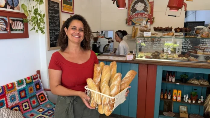 SC atinge 490 mil empresas lideradas por mulheres Uma foto de plano médio de Leslie Araújo, uma mulher com cabelos cacheados e vestindo uma blusa vermelha de decote ombro a ombro, sorrindo amplamente e segurando com as duas mãos uma cesta de arame branca cheia de várias baguetes douradas e artesanais de pão. Ela está em pé no interior de uma padaria aconchegante e rústica, com um balcão de serviço azul e madeira ao fundo. Atrás do balcão, outra mulher com um lenço na cabeça trabalha perto de uma vitrine de vidro cheia de doces e pães. Uma lousa de menu escrita à mão em português e uma placa redonda ilustrada que diz "Pão da Leli" são visíveis acima do balcão. À esquerda, prateleiras de madeira com fotos emolduradas de pães e plantas penduradas, e um banco de madeira com uma colcha de crochê colorida e de estilo "granny square". Uma prateleira azul mais baixa contém garrafas de azeite e outras embalagens. O ambiente é acolhedor e artesanal.