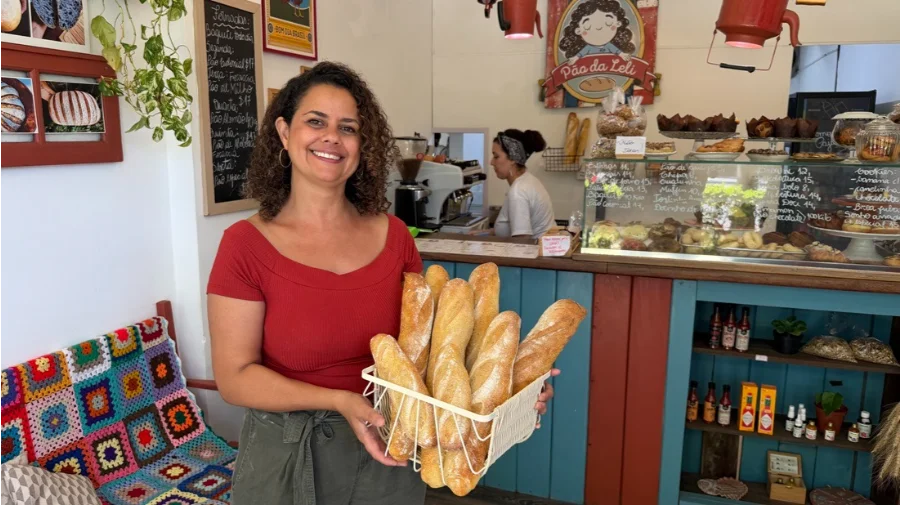 Uma foto de plano médio de Leslie Araújo, uma mulher com cabelos cacheados e vestindo uma blusa vermelha de decote ombro a ombro, sorrindo amplamente e segurando com as duas mãos uma cesta de arame branca cheia de várias baguetes douradas e artesanais de pão. Ela está em pé no interior de uma padaria aconchegante e rústica, com um balcão de serviço azul e madeira ao fundo. Atrás do balcão, outra mulher com um lenço na cabeça trabalha perto de uma vitrine de vidro cheia de doces e pães. Uma lousa de menu escrita à mão em português e uma placa redonda ilustrada que diz 