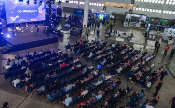 SC discute inovação pública no BrasilGov Summit 2026 Uma visão aérea panorâmica de um auditório de conferência em um grande salão de exposições. No canto superior esquerdo, um palco elevado tem um grande painel de LED exibindo "BRASILGOV SUMMIT". No palco, quatro painelistas estão sentados em poltronas modernas, e um apresentador de pé se dirige ao público. Uma grande audiência de centenas de pessoas está sentada em fileiras ordenadas de cadeiras azuis, voltadas para o palco. O fundo do salão mostra uma área de exposição com vários estandes de empresas (como "unifique" e "wacon") e pessoas circulando. A iluminação do local é natural e de palco, criando uma atmosfera ativa de evento.